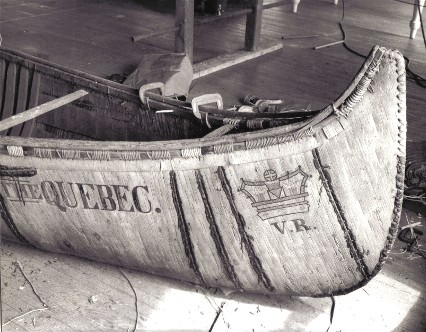 Bow view of  25' fur-trade type birch bark canoe , ''THE QUEBEC'', under repair by Henri Vaillancourt and Rick Nash in 1972. It features the royal crown painted on both ends with the initials V.R. [ Victoria Regina ] underneath.