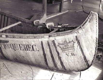 Bow view of 25' fur-trade type birchbark canoe , ''THE QUEBEC'', under repair by Henri Vaillancourt and Rick Nash in 1972. It features the royal crown painted on the ends with the initials V.R. [ Victoria Regina ] underneath.