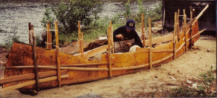 Basil Smith uses split spruce root to sew the seams of a new birchbark canoe . The birchbark  has been laid out on a sand building bed and staked into the shape of the canoe ; from '' Building an Algonquin Birchbark Canoe '' ; photo Henri Vaillancourt , Maniwaki , Quebec 1980